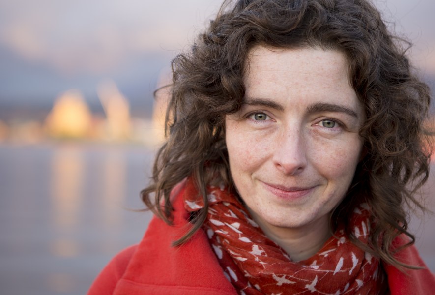 A photo portrait of a smiling woman with medium length dark curly hair. She is standing in front of a shoreline with water in the background. 